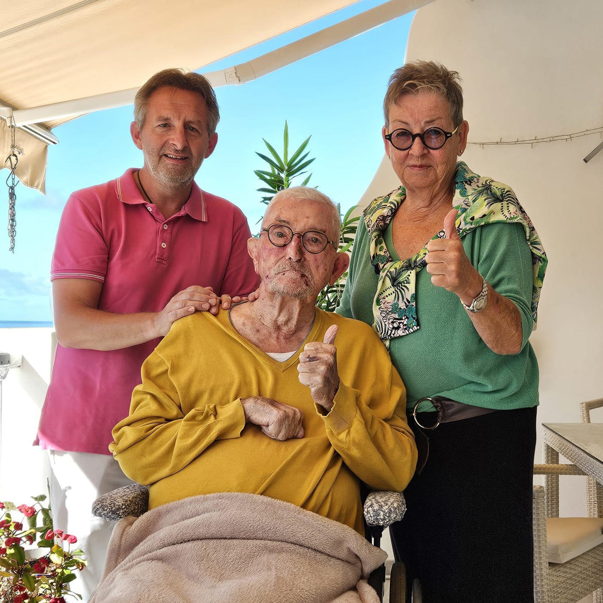 Bernd Weisser im rosafarbenen Poloshirt sitzt neben einem älteren Herrn auf einer Terrasse am Meer. Beide blicken freundlich in die Kamera.